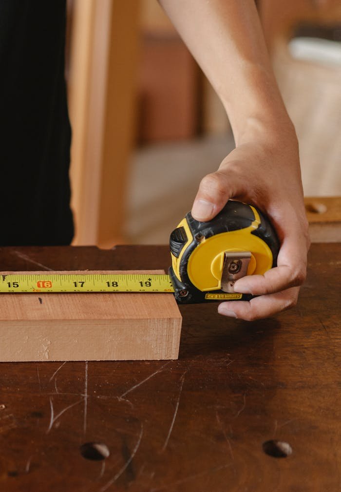 Unrecognizable male carpenter using measuring tape on wooden board while working at scratched workbench in professional joinery on blurred background